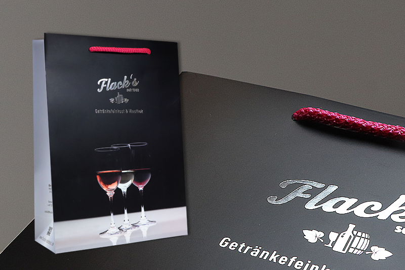 Papiertasche Flack's GetränkeFeinkost & Vinothek glänzend geprägt mit silber auf schwarz-grauem Grund, pinkfarbige Kordel, Detailaufnahme der Prägung