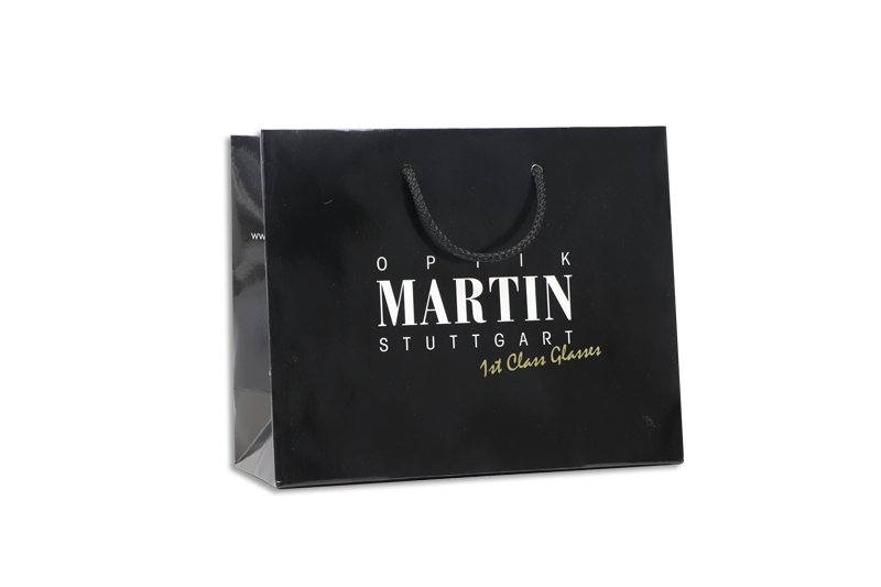 Schwarze, glänzende Papiertasche mit Werbeaufdruck Optik Martin in weiß und gold, schwarze Tragekordeln
