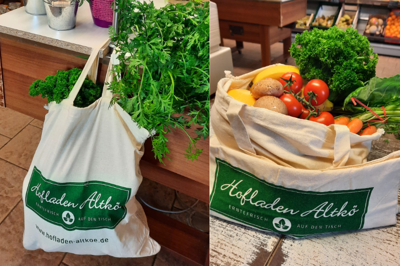 Zwei naturfarbige Baumwolltaschen grün bedruckt mit Werbung für den Hofladen Altkö. Beide Taschen sind mit Obst und Gemüse befüllt. Produktbild für Tausendtypentragetaschen